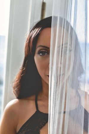 Woman in black attire poses near window with sheer curtains in natural light.