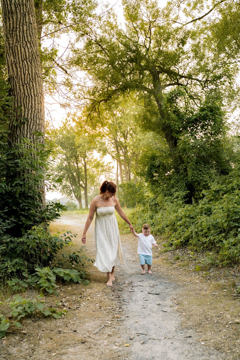 A scenic wooded path with dappled sunlight filtering through trees as two figures walk together in white clothing.