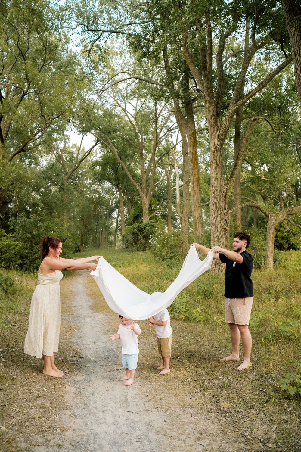 A sequence of playful moments on a tree-lined dirt path shows a family enjoying a summer day with a white blanket.