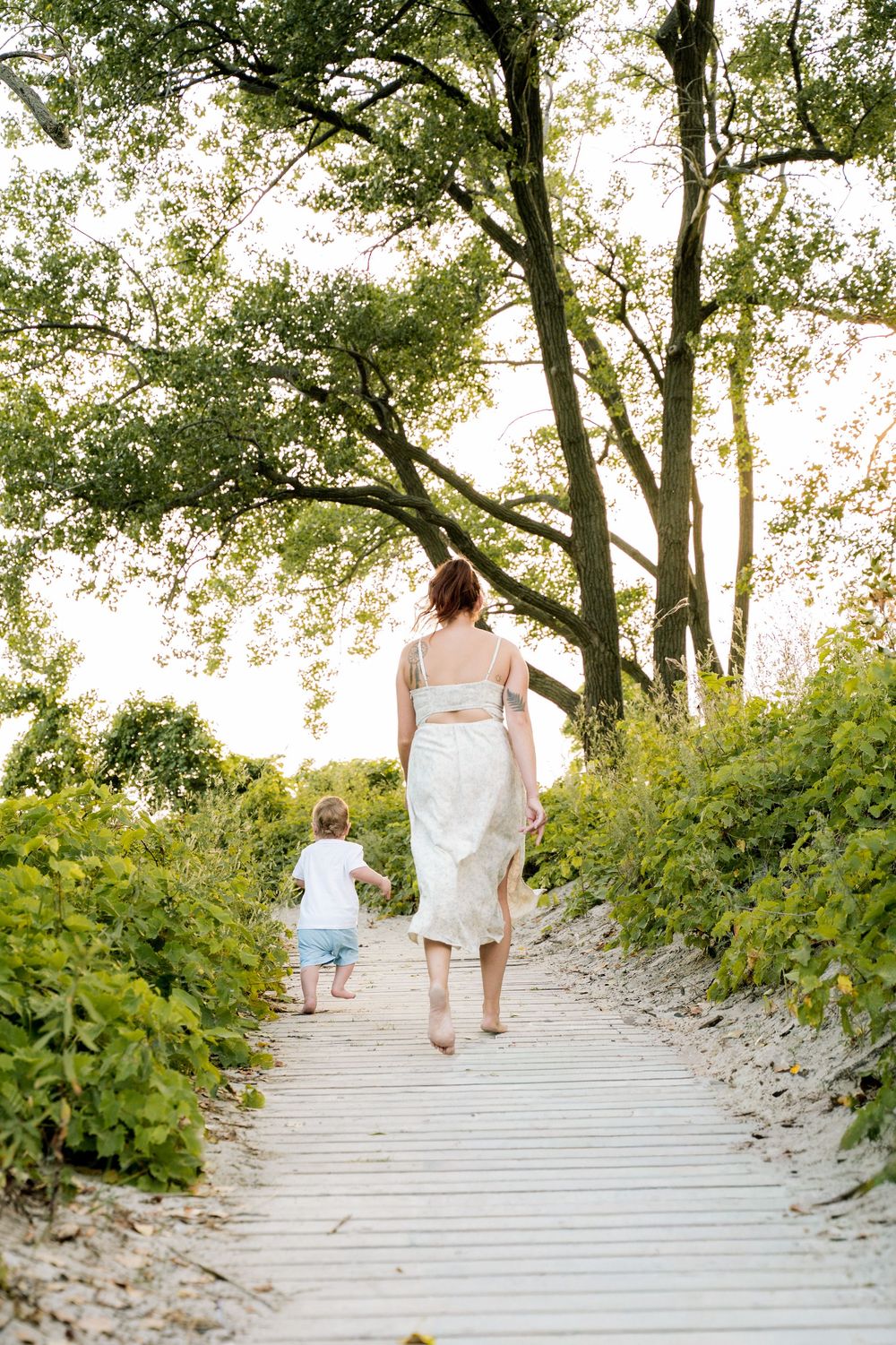 A couple in white outfits walks up wooden stairs surrounded by lush greenery on a sunny day.