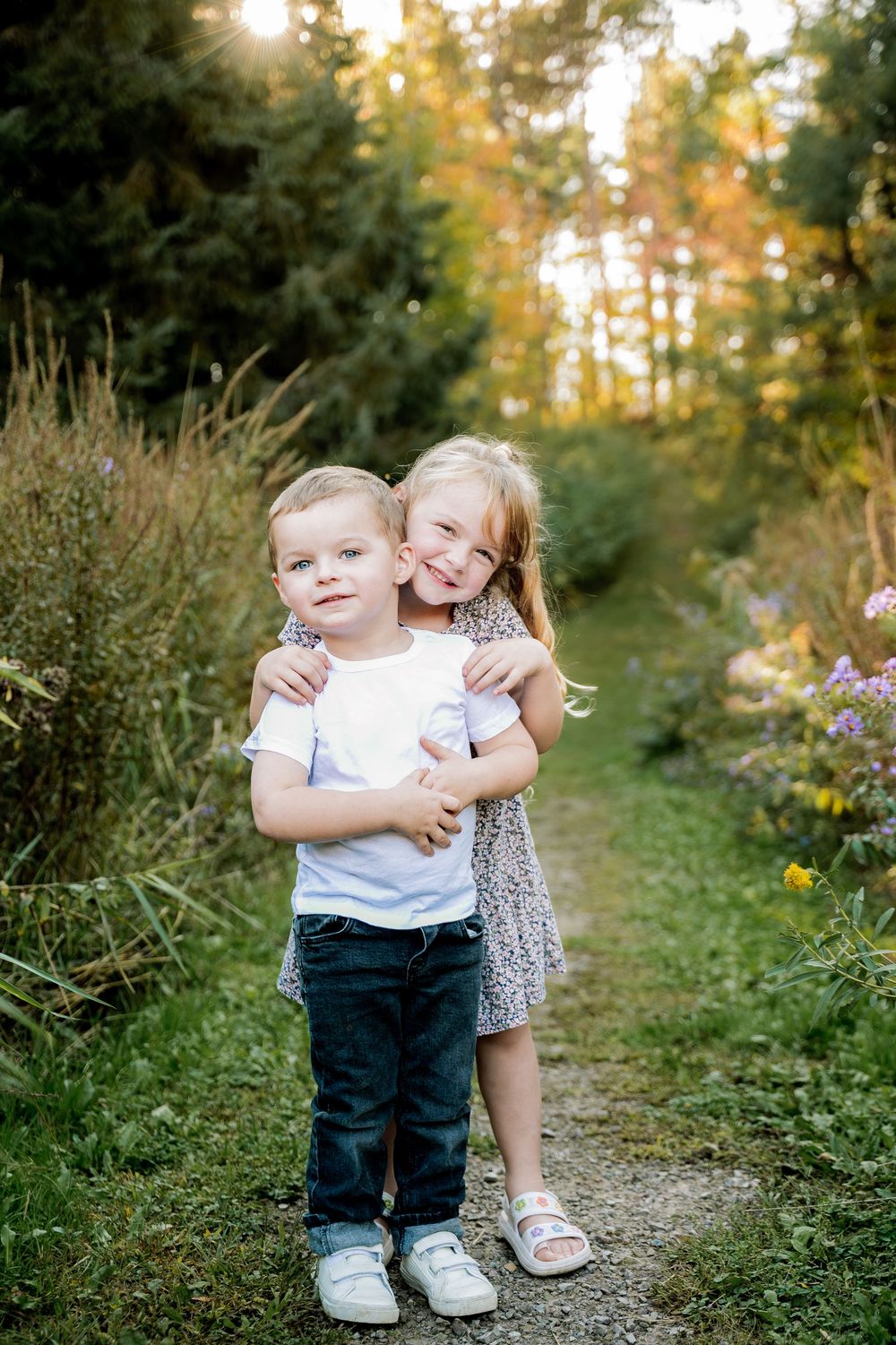 Two young children share a playful moment on a nature path surrounded by wildflowers at sunset.