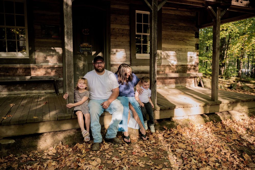 A family sits together on the wooden steps of a rustic cabin porch surrounded by autumn leaves.