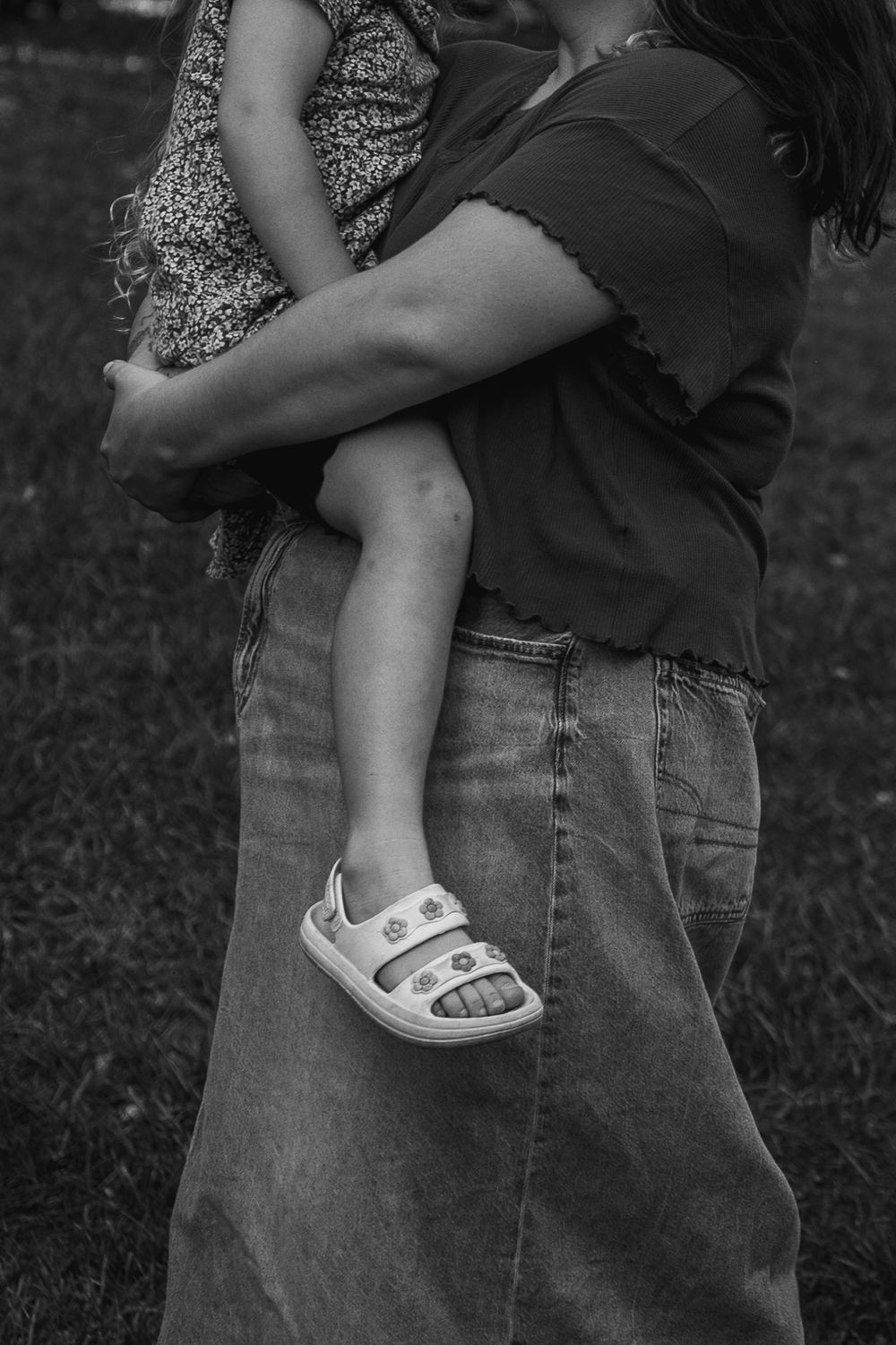 Black and white photo of a child wearing sandals being held up against someone in jeans in an outdoor setting.