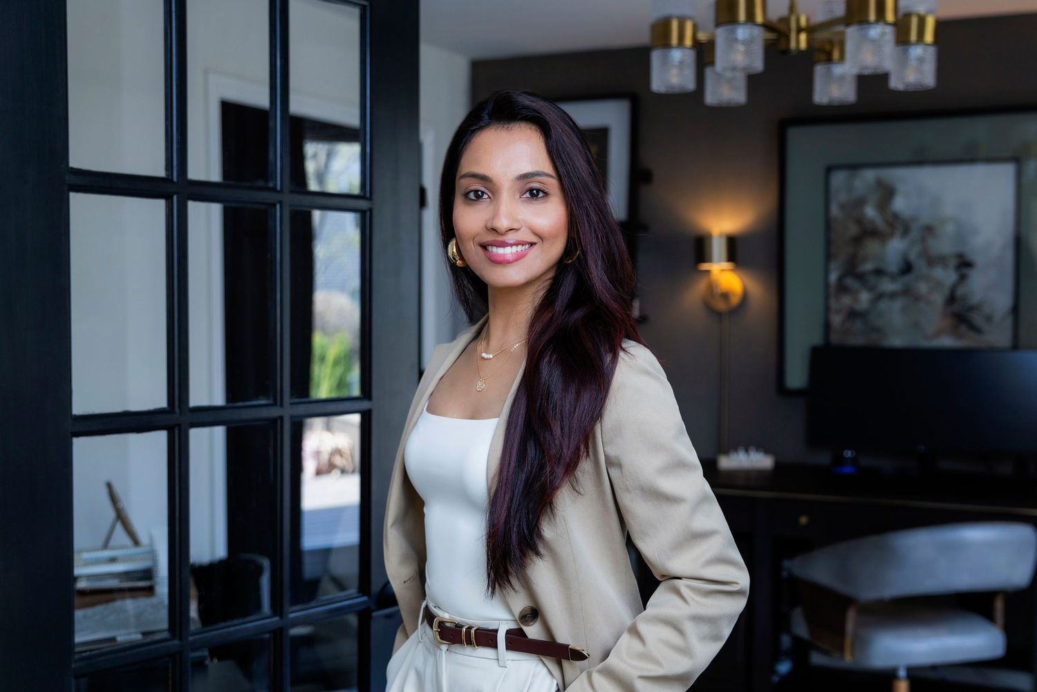 Professional in a beige blazer smiles while standing in a modern office setting with dark walls and warm lighting.
