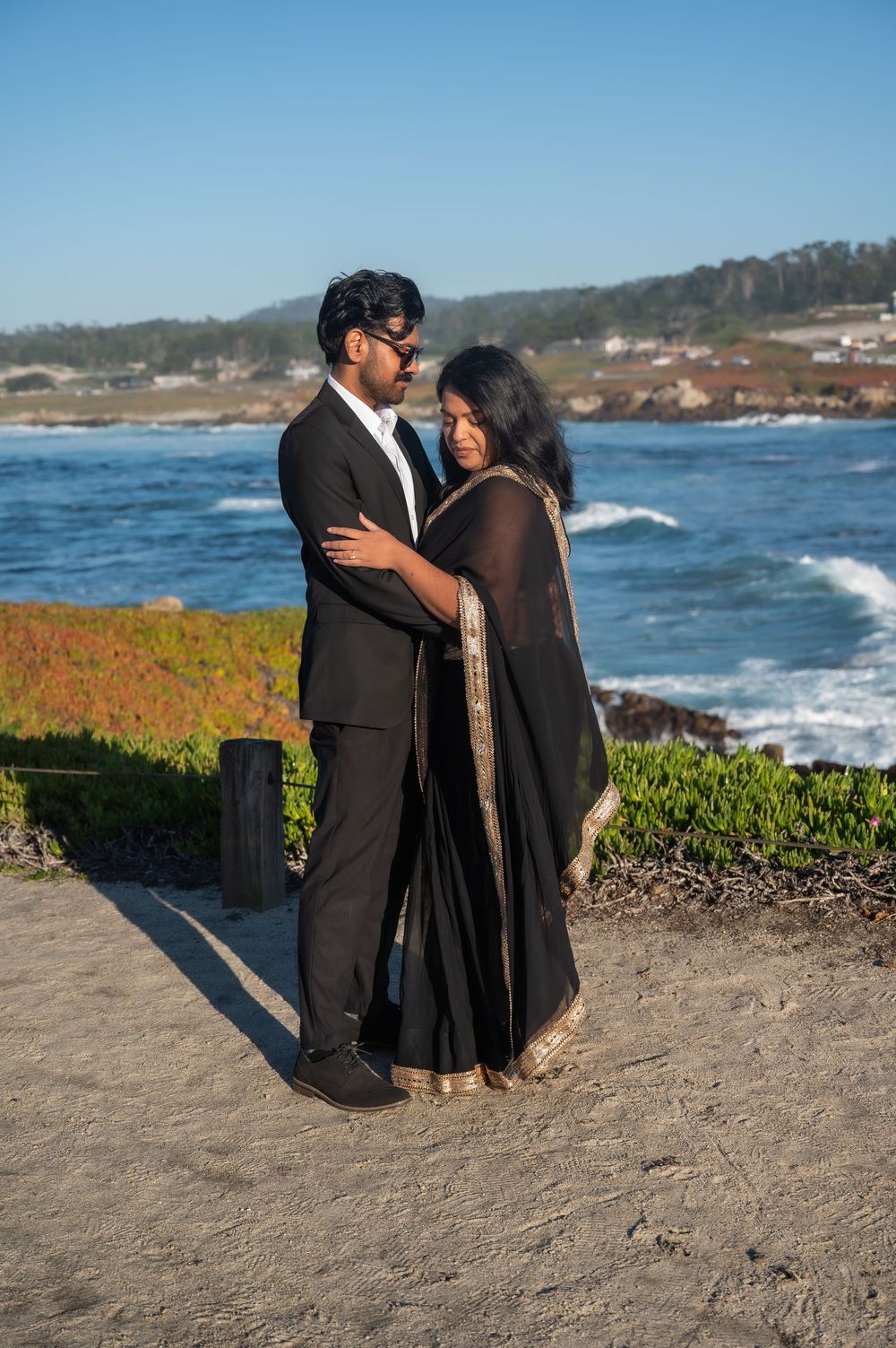 A couple in formal attire embraces on a scenic coastal cliff overlooking the ocean waves and shoreline.