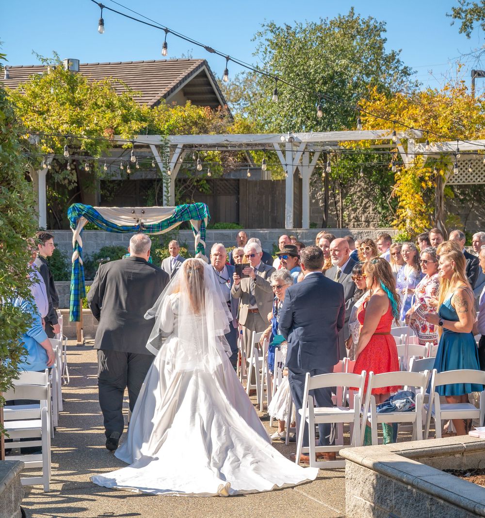 A beautiful outdoor wedding ceremony setup with white chairs and guests gathered under string lights and trees.