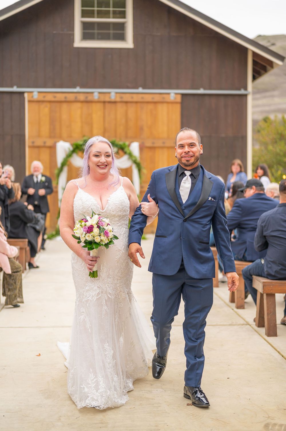 A happy couple walks together on a rustic path outside a barn venue during their wedding celebration.