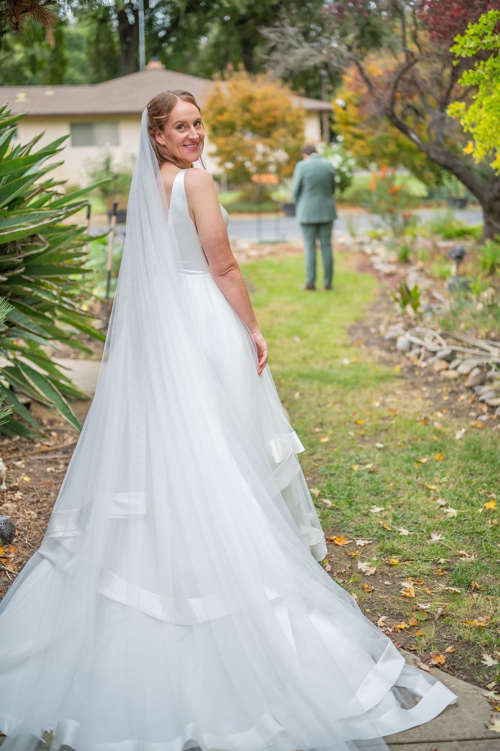 A bride in a flowing white layered wedding gown with a long veil poses in a garden setting during autumn.