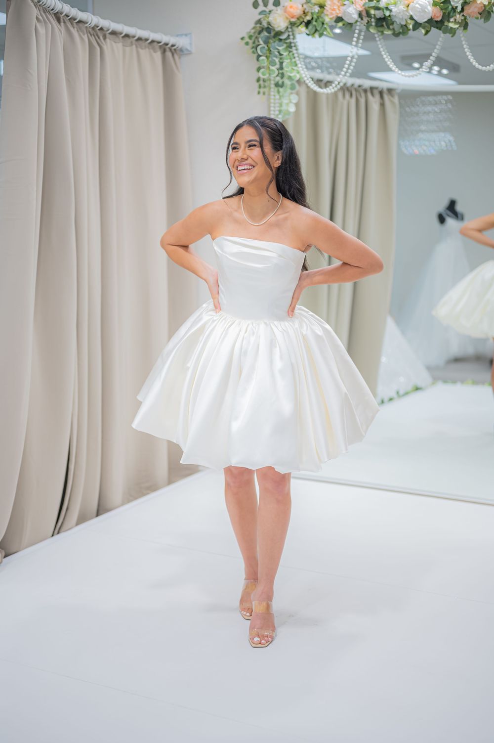 Woman in white strapless mini dress twirls playfully in a light-filled room with floral decorations.