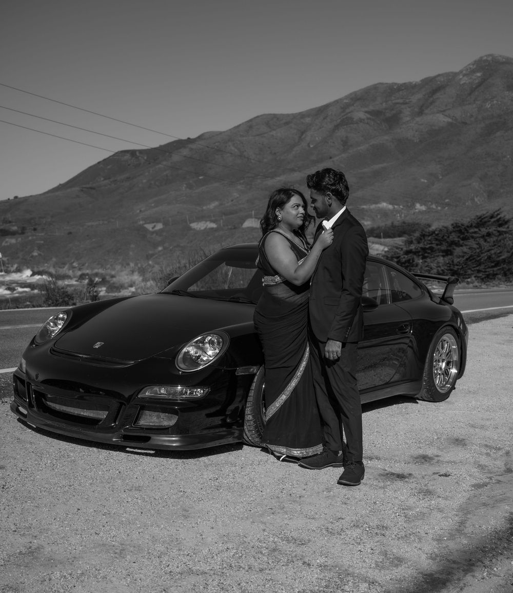 A couple poses romantically against a black Porsche 911 with mountains in the background in black and white.