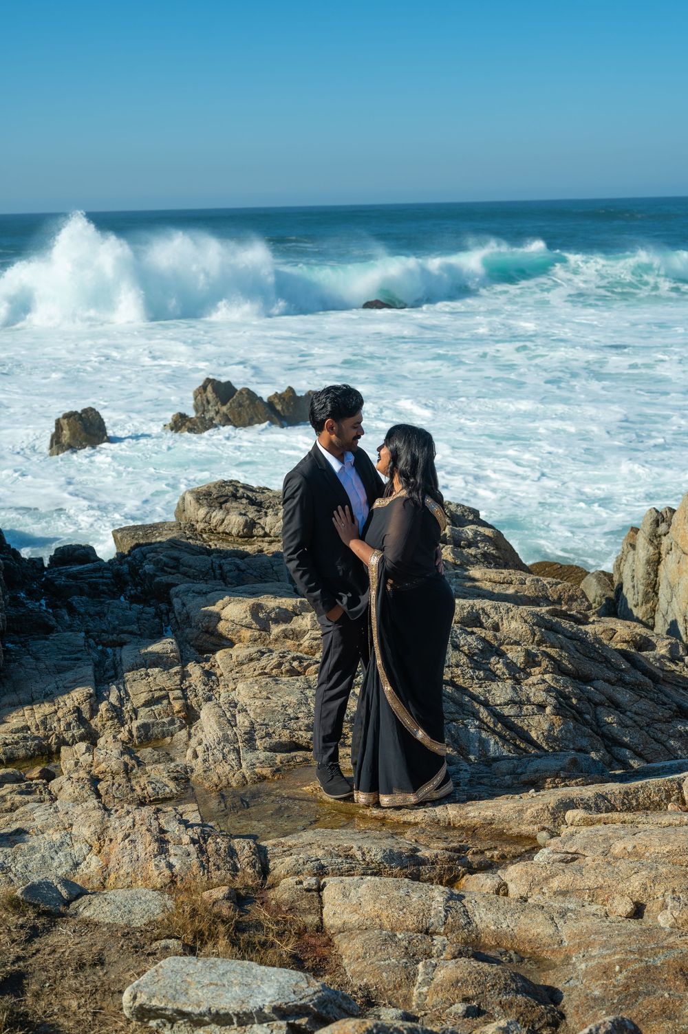 A photographer captures dramatic ocean waves crashing against rocky coastal cliffs on a sunny day.