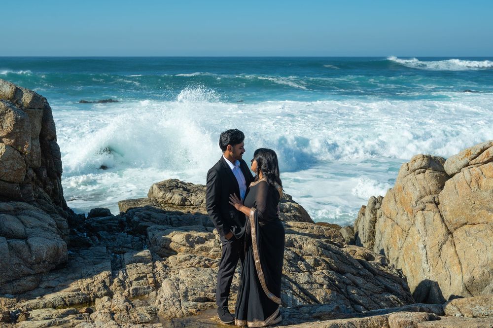 A romantic couple embraces on rocky coastal cliffs with crashing ocean waves in the background.