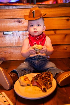 A toddler in a cowboy hat and red bandana sits at a wooden table with a plate of steak and french fries.