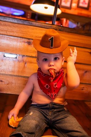 A baby in western-style attire with jeans, red bandana and brown baseball cap sits on wooden benches.