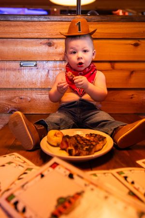 A small diner patron in a cowboy hat sits at a wooden table with a plate of barbecue food.