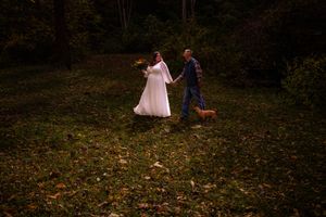 Couple in formal attire walk together through autumn woods during golden hour photoshoot.