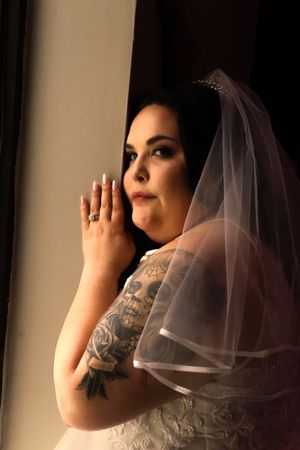 Bride in white wedding dress with visible tattoos and veil poses in dramatic side lighting.
