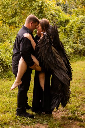 A couple embraces romantically outdoors while one wears dramatic black feathered wings as a costume piece.
