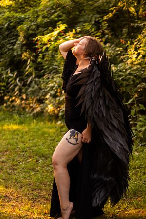 A figure in a dramatic black dress with feathered wings poses dramatically against autumn foliage at sunset.