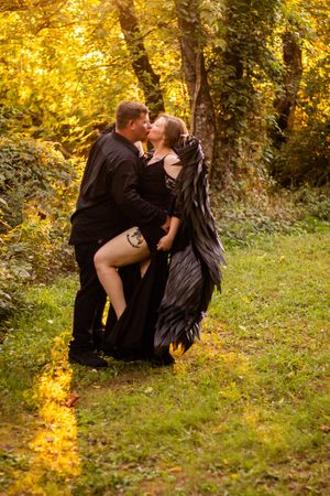 A romantic couple in black attire embrace in a sunlit autumn forest with golden foliage.