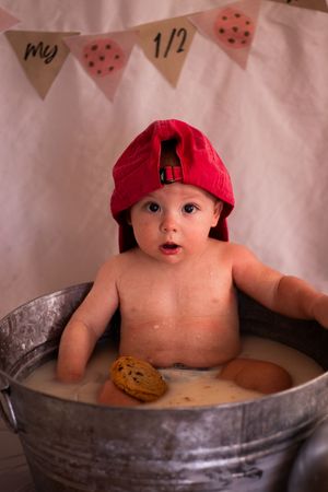 A baby wearing a red cap sits in a metal basin with a cookie or pastry.