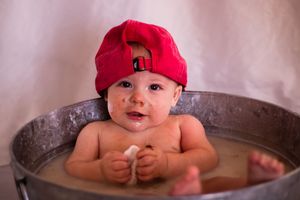 Baby wearing red hat smiles while sitting in wooden bathtub filled with soapy water.