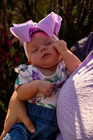 Baby wearing floral outfit and purple bow lying on grass with dreamy expression in outdoor portrait series.