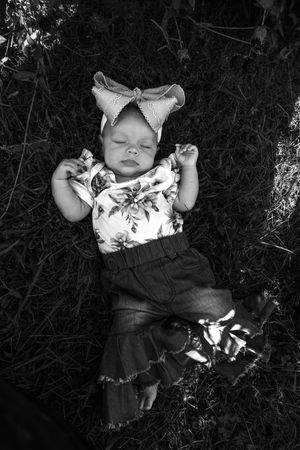 A baby wearing a floral dress lies peacefully on dark grass in a black and white artistic photograph.