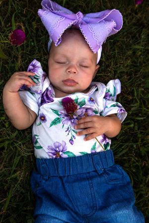 Infant in denim and floral print outfit resting peacefully on grass with purple bow accessory.