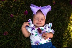 A baby wearing a purple bow and floral outfit lies on grass in sunlight with a joyful expression.