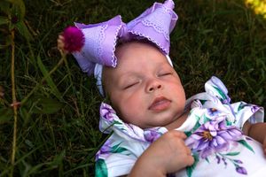 Baby in white floral dress and purple bow relaxing outdoors in garden setting.