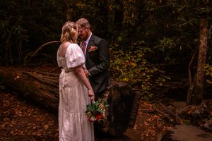 A couple in vintage-style white dresses poses romantically in a moody autumn forest setting with fallen leaves.