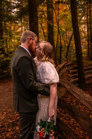 A romantic kiss shared between newlyweds in a forest setting with autumn leaves and fallen logs.
