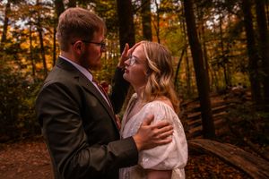 A romantic couple in formal attire embraces during a sunset photoshoot in an autumn forest setting.