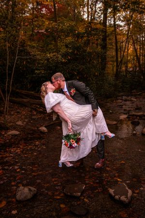 Romantic autumn wedding couple poses in a wooded setting with fallen leaves and rocks at sunset.