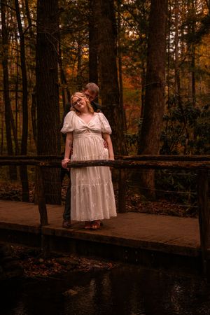 A figure in a white vintage dress poses on a wooden boardwalk in an autumn forest at dusk.