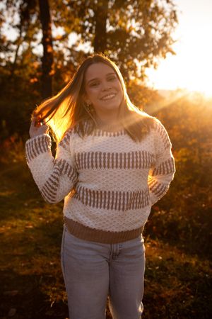 Person in white crochet sweater poses outdoors at sunset with golden sunlight filtering through trees.