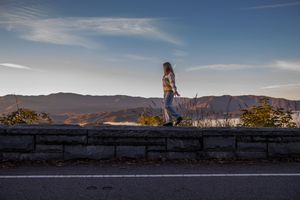 A silhouetted figure sits on a stone wall at sunset against a mountain landscape backdrop.