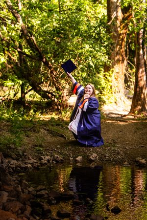 A person in blue graduation attire sits contemplatively by a tranquil forest pond surrounded by lush greenery.
