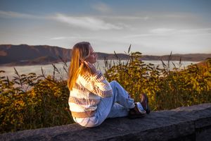 A person in a denim jacket sits on a stone wall at sunset overlooking a scenic mountain landscape.