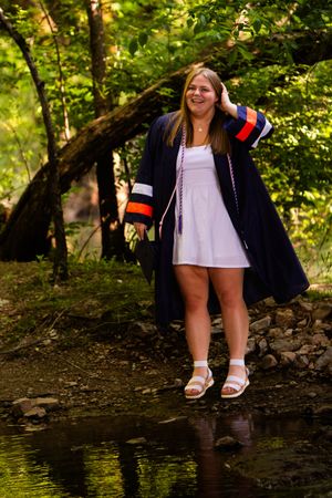 A person in a navy cardigan and white dress stands near a reflective pond in a wooded setting.