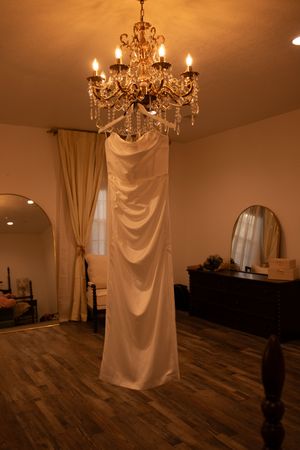 An elegant white wedding dress hangs beneath a crystal chandelier in a room with hardwood floors and soft lighting.
