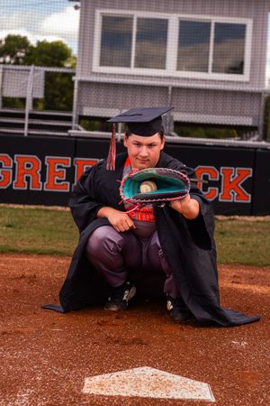 A graduate in cap and gown poses playfully sliding into home plate on a baseball diamond.