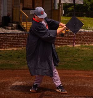 A graduate in black cap and gown poses with their diploma on a dirt field near brick buildings.