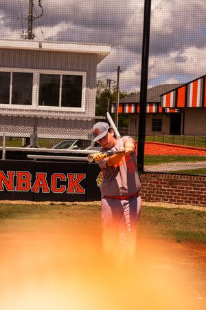 Baseball player in orange uniform swings bat during practice at field with brick building and cloudy sky in background.