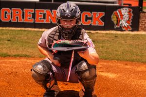 Baseball catcher in protective gear crouches on orange clay dirt behind home plate during a game.