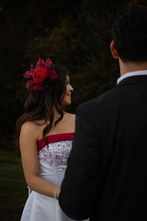 A couple in wedding attire stands close together outdoors at dusk, with the bride wearing a white dress and red floral hairpiece.