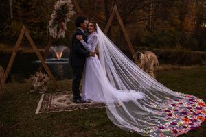 Long white wedding veil with colorful floral embroidery trailing behind couple in outdoor setting.