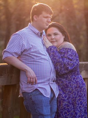 A couple embraces against a rustic wooden fence at sunset, with the woman wearing a purple floral dress.