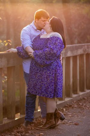 A couple shares a romantic moment at sunset against a wooden fence with autumn leaves and purple flowers in view.